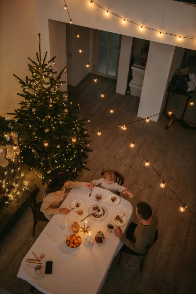 winter traditions for families. Image of mother, father, and child holding hands around dinner table with candles and cake. Image has Christmas tree in the background and string lights.
