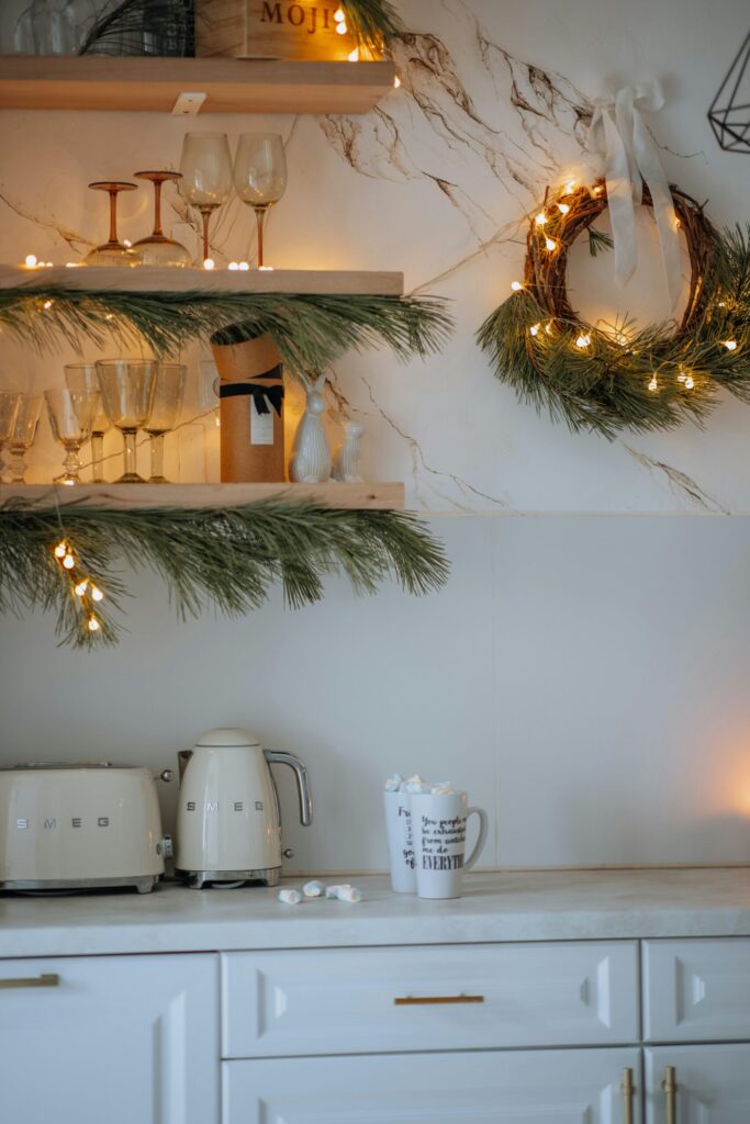 ideas from winter traditions. Image of counter with winter decorations (garland and lights). There are wooden bookshelves with glasses, white counter with white Smeg toaster and kettle on white counter with white cabinets. 