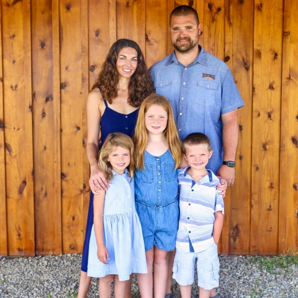 mom of three. Image of family of 5 (mom dad, two girls, and one boy) wearing blue and standing in front of a wooden barn.