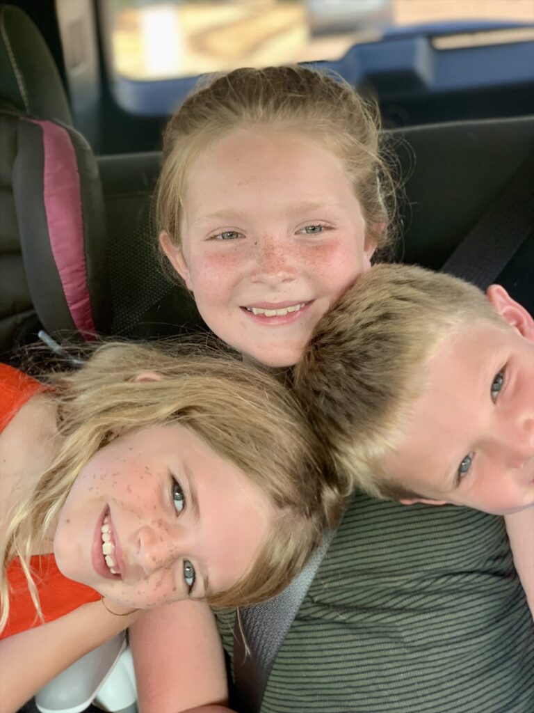 real motherhood. Image of three children (two girls and one boy) in a car. The girl is sitting in the middle and a boy and girl are leaning with their heads on each of her shoulders.