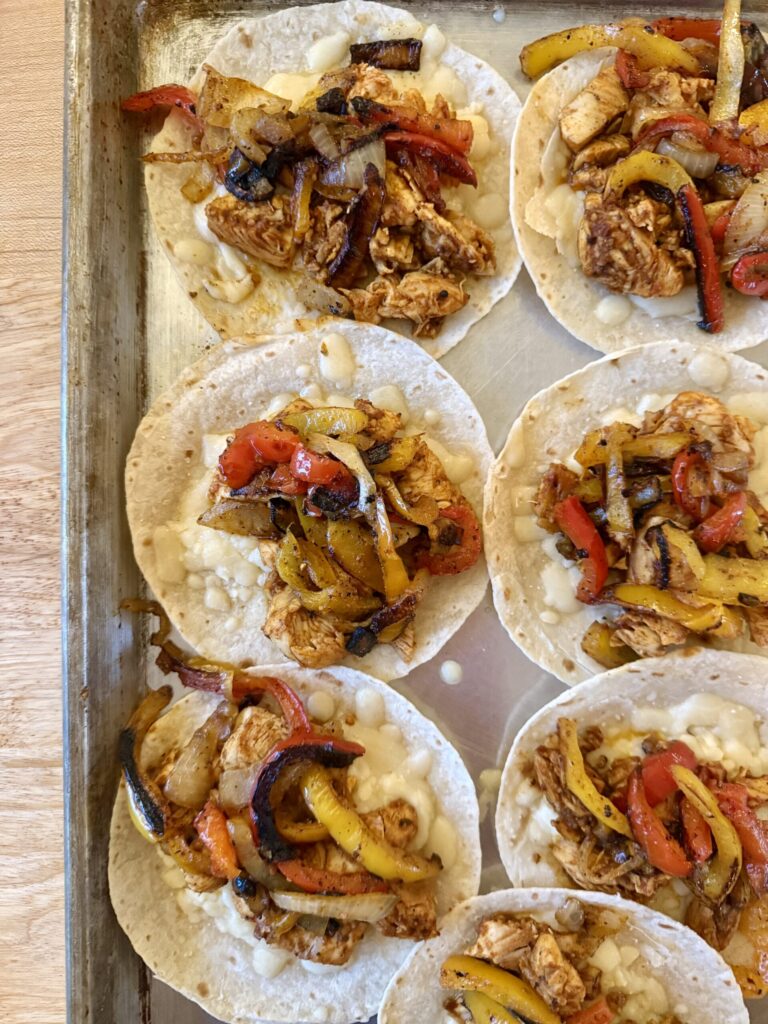 sheet pan tacos with shredded chicken, sauteed bell peppers and onions, and cheese on sheet pan on wooden counter.