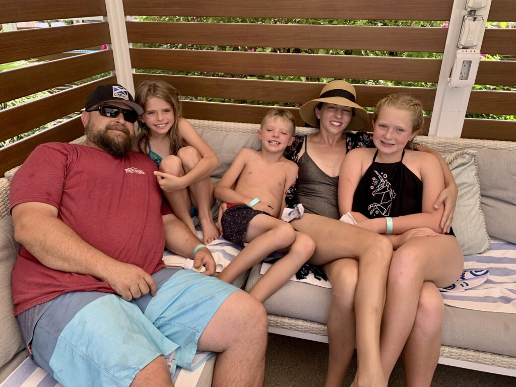 working mom. Picture of family - mom, dad, two daughters and a son in swimsuits sitting on outdoor furniture.