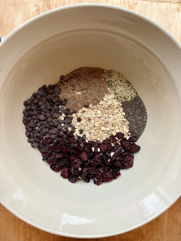 Image of ingredients for oatmeal protein balls (oats, chocolate chips, flax, hemp hearts, chia seed, dried cherries) in cream ceramic bowl. 